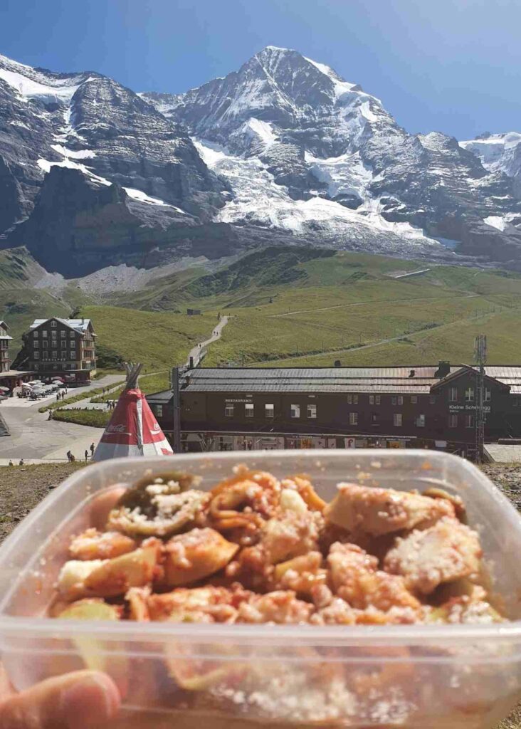 Un tupper con pasta frente a las montañas de los alpes suizos, cocinar es una forma de comer en suiza barato